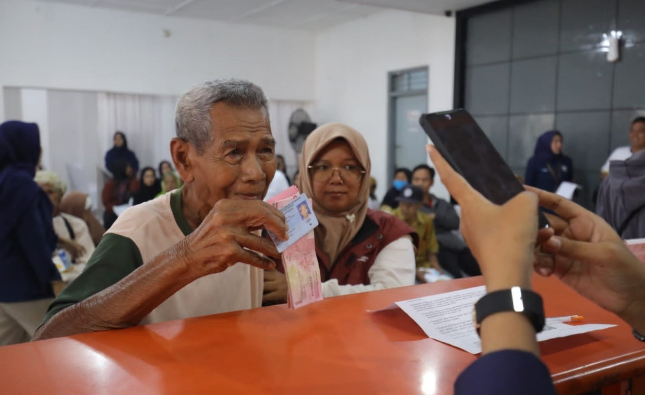 <p>Ilustrasi seorang warga lanjut usia menunjukkan KTP dan buku tabungan saat proses pencairan bantuan sosial di kantor desa. Pemerintah kini memprioritaskan 2 juta warga miskin baru sebagai penerima PBI JKN 2025, menyusul penyisiran data bantuan yang tidak tepat sasaran. Foto: Dok. kemensos.go.id</p>
